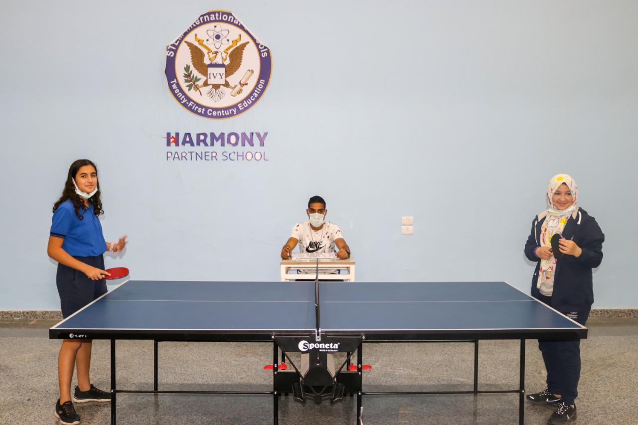 Students playing table tennis at IVY STEM International School, with a young girl ready to strike the ball, another student waiting her turn, and an observer wearing a face mask.